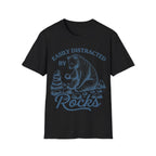 Linocut-style illustration of a bear sitting down examining a stone beside a cairn stack, framed by text reading 'EASILY DISTRACTED BY ROCKS'.
