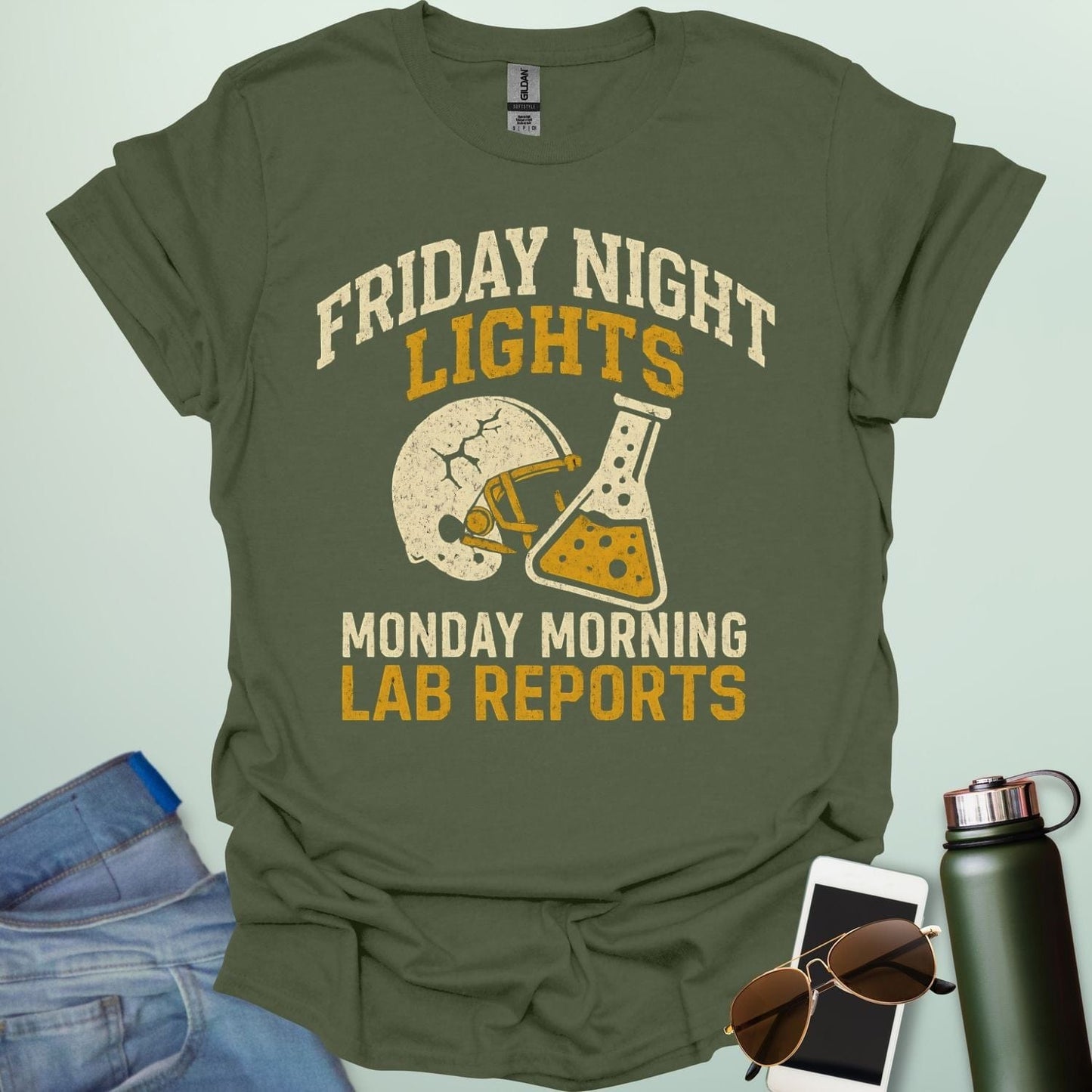 A military green t-shirt with a vintage football helmet and a cracked lab flask, with the text 'FRIDAY NIGHT LIGHTS' and 'MONDAY MORNING LAB REPORTS.'A t-shirt with a vintage football helmet and a cracked lab flask, with the text 'FRIDAY NIGHT LIGHTS' and 'MONDAY MORNING LAB REPORTS.'
